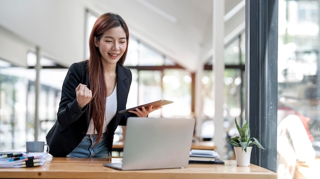 Elegante Geschäftsfrau, die im Büro mit digitalem Tablet steht Aufgeregte asiatische Geschäftsfrau, die die Hände hebt, um zu gratulieren, während sie am Laptop im Büro arbeitet