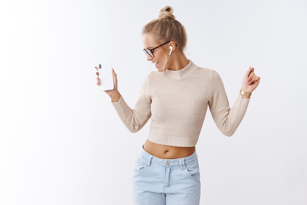 Elegante glamour y despreocupada mujer caucásica en gafas de suéter recortadas alejándose alegremente agitando las manos en la danza sosteniendo el teléfono inteligente escuchando música en auriculares inalámbricos sobre fondo blanco.