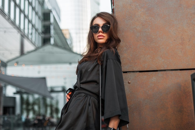 Elegante glamour hermosa dama urbana elegante con elegantes gafas de sol y un traje de noche negro de moda con un bolso cerca de la pared en la ciudad Mujer bonita