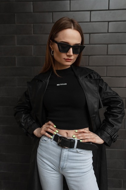 Elegante y glamurosa modelo de mujer bonita joven vestida de negro a la moda con un abrigo de cuero largo a la moda con una camiseta negra y jeans vintage de pie cerca de una pared de ladrillo negro