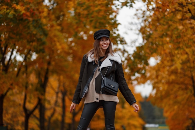 Elegante glückliche junge Frau in einem schicken Hut in einer stilvollen Jacke mit einer Tasche im Wald auf einem Hintergrund der goldenen Blätter. Fröhliches Mädchen geht in den Park. Herbstkollektion trendiger Damenbekleidung.