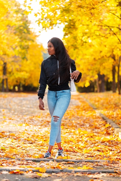 Elegante hermosa joven afroamericana en ropa de moda con una chaqueta negra y jeans con un bolso camina en un parque de otoño con follaje de otoño de color amarillo