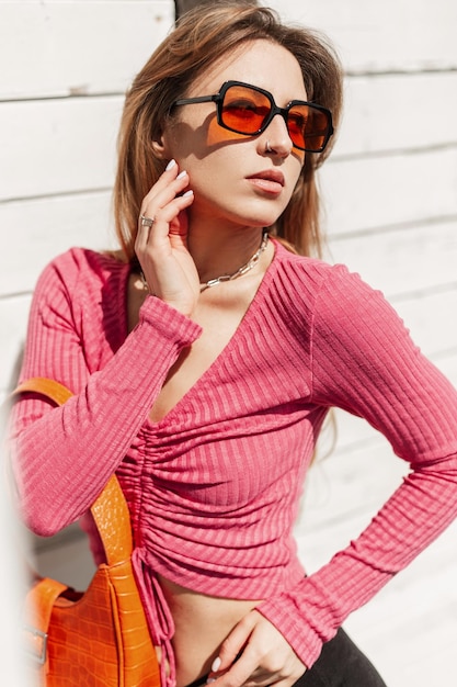 Foto elegante hermosa joven pelirroja con gafas de sol naranjas de moda en ropa de verano brillante de moda con una bolsa de cuero posa cerca de un edificio de madera blanca en la playa