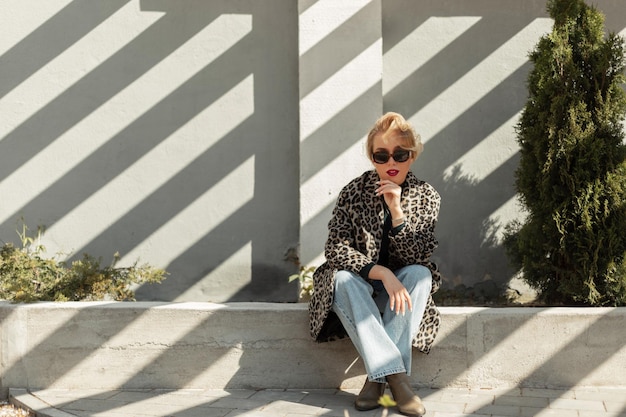 Elegante hermosa joven rubia con gafas de sol en un abrigo de leopardo de moda con jeans acampanados y zapatos se sienta en la calle con luz solar