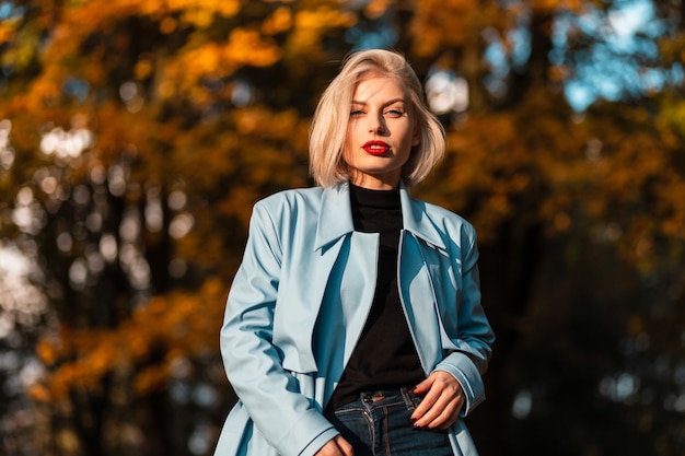 Elegante hermosa joven rubia con labios rojos en un abrigo azul camina en un parque de otoño con follaje de otoño naranja al atardecer