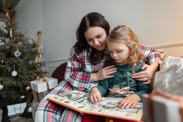 Elegante y hermosa madre e hija se sientan en el sofá con regalos cerca del árbol de Navidad y leen un libro en la víspera de Navidad en casa