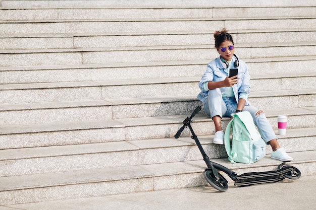 Foto elegante y hermosa mujer joven sentada en los pasos después de montar en scooter eléctrico y enviar mensajes de texto a amigos o consultar las redes sociales