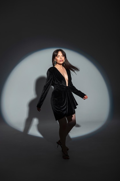 Elegante hermosa mujer modelo chic con peinado en un vestido negro de moda con un escote posando en movimiento en el estudio en la oscuridad con luz circular