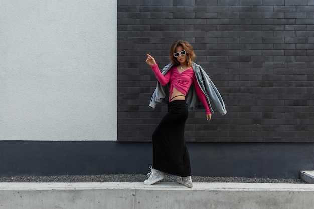 Elegante y hermosa mujer pelirroja hipster de moda con gafas de sol blancas frescas con ropa brillante de moda con chaqueta de mezclilla de moda falda rosa y zapatillas bailando cerca de la pared en la calle