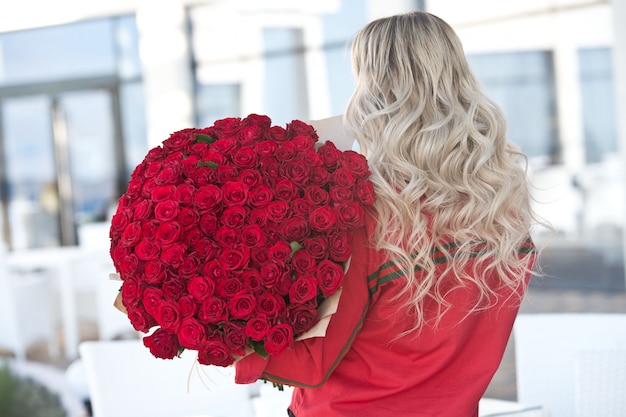 Elegante hermosa mujer rubia lleva vestido de moda verde en café con gran ramo de 101 rosas rojas.