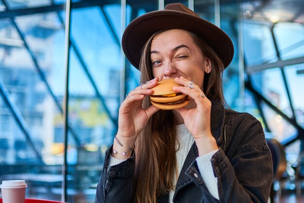 Elegante hipster hambrienta come pan de hamburguesa con carne en restaurante de comida rápida.