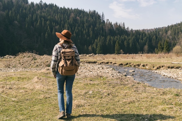 Elegante hipster con sombrero marrón y suéter de lana con mochila textil vintage cerca de montañas, ríos y bosques Mujer de cabello rubio explorando la naturaleza Concepto de viaje y pasión por los viajes Increíble atmósfera