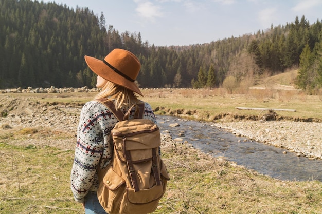 Elegante hipster con sombrero marrón y suéter de lana con mochila textil vintage cerca de montañas, ríos y bosques Mujer de cabello rubio explorando la naturaleza Concepto de viaje y pasión por los viajes Increíble atmósfera