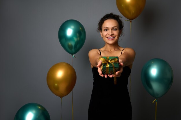 Elegante hispanische Frau im schwarzen Abendkleid zeigt ein Weihnachtsgeschenk in glänzendem Packpapier und goldener Schleife, lächelt zahniges Lächeln in die Kamera. Grauer Hintergrund mit goldgrünen metallischen Ballons