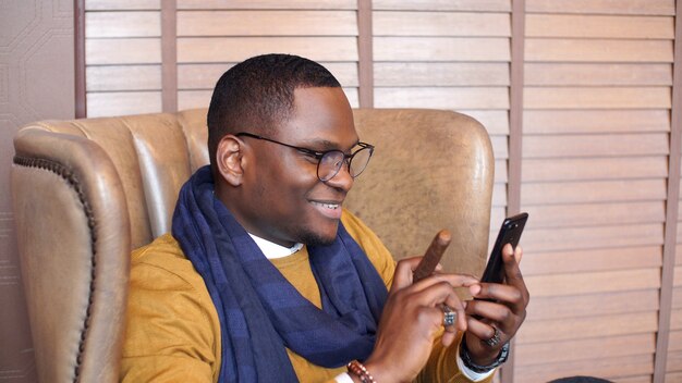 Elegante hombre afroamericano feliz está sentado en una silla con un cigarro