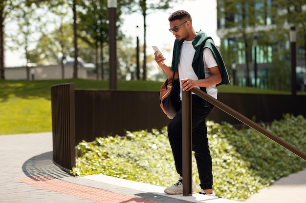 Foto elegante hombre afroamericano con gafas de sol sosteniendo un teléfono inteligente mientras camina en el área urbana enviando mensajes de texto o usando el navegador