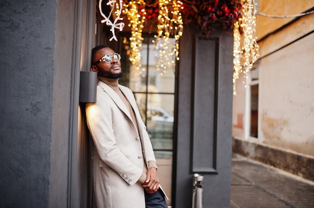 Elegante hombre afroamericano usa chaqueta beige con gafas de sol posan contra la casa con guirnalda dorada.