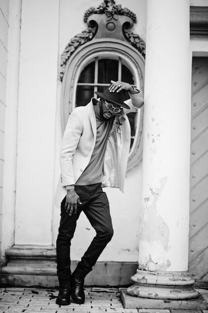 Elegante hombre afroamericano usa chaqueta beige y sombrero negro con gafas de sol posan contra la ventana vintage.