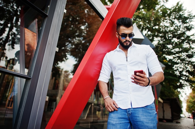 Elegante hombre alto con camisa blanca, jeans y gafas de sol posando en la calle de la ciudad Barba chico atractivo mirando el teléfono celular rojo.