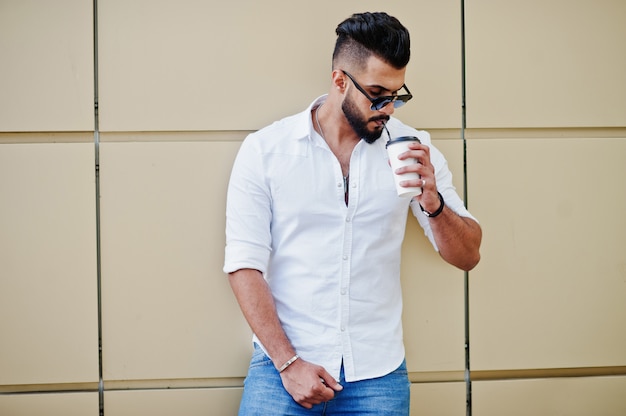 Elegante hombre alto con camisa blanca, jeans y gafas de sol posando en la calle de la ciudad