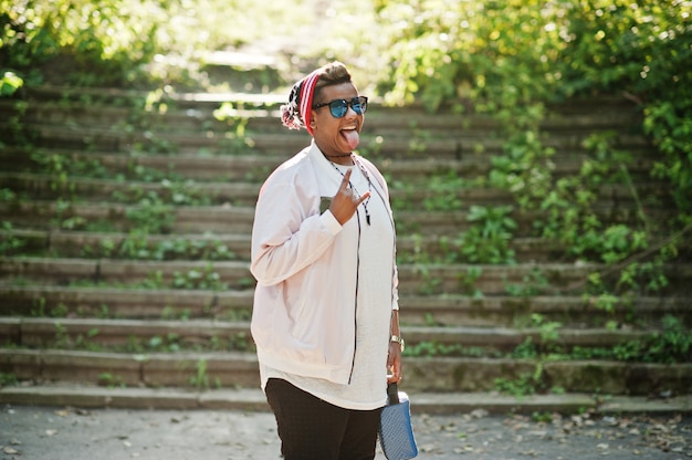 Elegante hombre árabe hipster hombre con sombrero y gafas de sol posando al aire libre en la calle, muestra tonque y dos dedos.