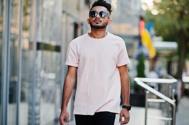 Elegante hombre de barba india con gafas de sol y camiseta rosa Modelo de India posado al aire libre en las calles de la ciudad