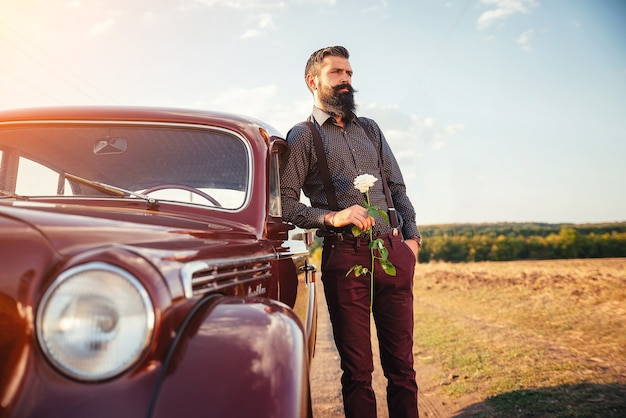 Elegante hombre barbudo con bigote en pantalones clásicos con tirantes y una camisa oscura con una rosa en sus manos en un coche retro marrón