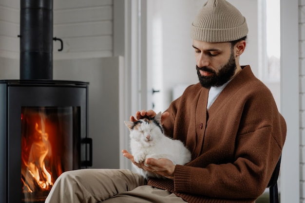 elegante hombre barbudo con su gato en casa sentado cerca de la chimenea