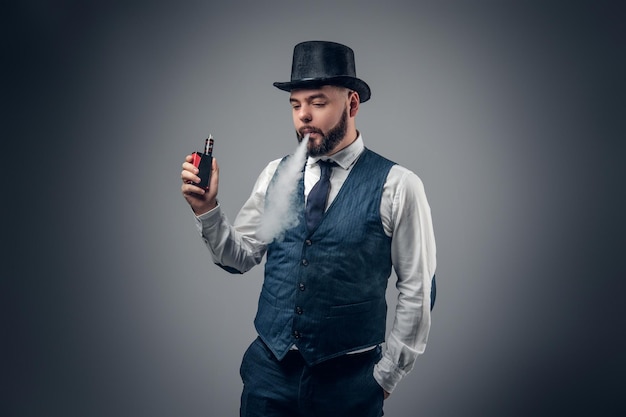 Un elegante hombre barbudo vestido con chaleco y sombrero de copa fumando un cigarrillo electrónico.