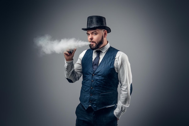 Un elegante hombre barbudo vestido con chaleco y sombrero de copa fumando un cigarrillo electrónico.