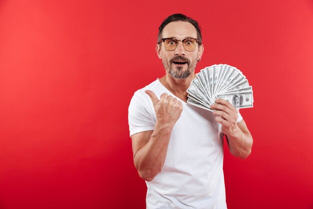 Elegante hombre caucásico de 30 años en camiseta blanca casual con gafas sosteniendo un montón de dinero en efectivo en dólares y apuntando con el dedo a un lado en copyspace, aislado sobre fondo rojo