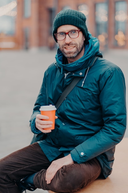 Elegante hombre caucásico tiene tiempo libre, posa afuera con una taza de café desechable