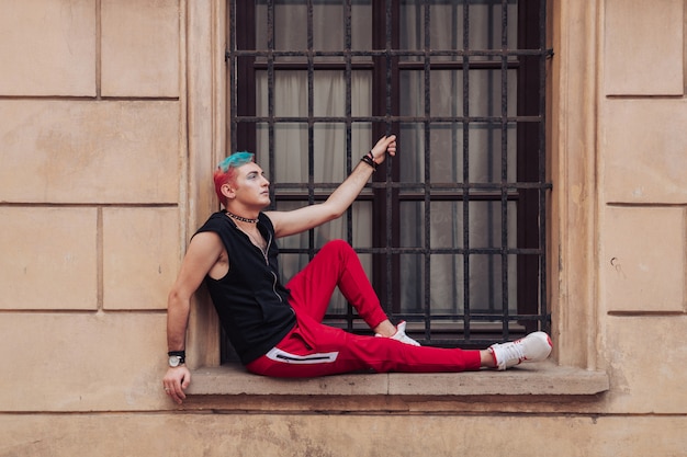 Elegante hombre homosexual con maquillaje, cabello colorido, collar de cuero con púas posando en la pared amarilla con ventana