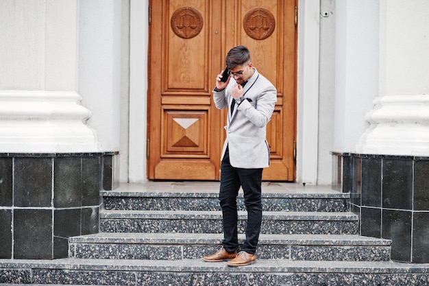 Elegante hombre indio con bindi en la frente y anteojos con traje gris posado al aire libre contra la puerta del edificio hablando por teléfono móvil