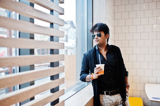 Elegante hombre indio en gafas de sol permanecer cerca de la ventana con una taza de café en la cafetería de comida rápida.