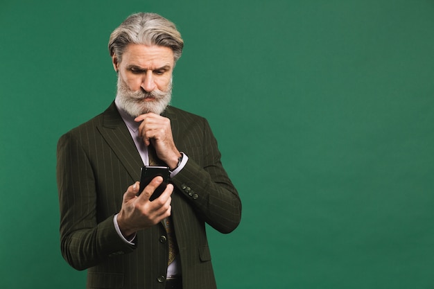 Elegante hombre de mediana edad en traje mira el teléfono y arregla el bigote y la barba en la pared verde