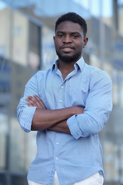 Elegante hombre de negocios afroamericano con los brazos cruzados en puestos de camisa azul y sonríe a la cámara