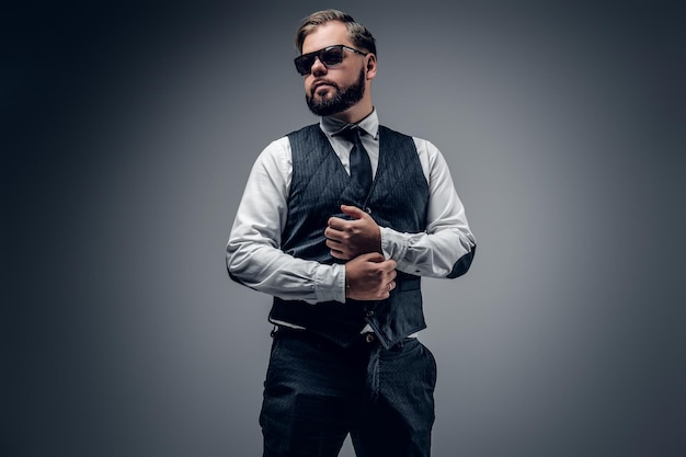 Un elegante hombre de negocios barbudo con gafas de sol y chaleco sobre fondo gris.