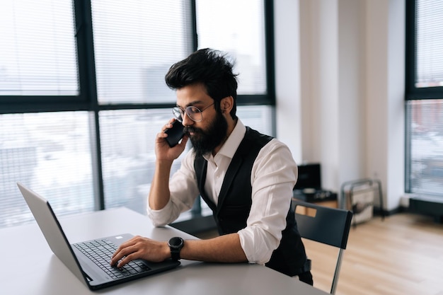 Elegante hombre de negocios indio barbudo con anteojos trabajando en una computadora portátil sentado en el escritorio y hablando por teléfono inteligente en el fondo de la ventana