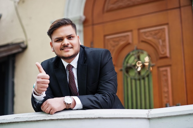 Elegante hombre de negocios indio con ropa formal apoyado en una barandilla contra la puerta en el centro de negocios Él es un hombre feliz y muestra el pulgar hacia arriba