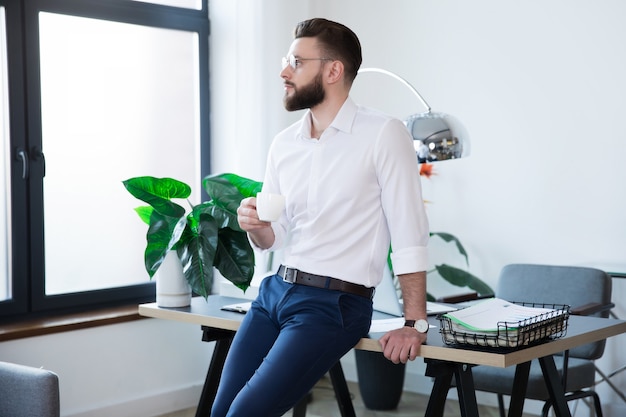 Elegante hombre de negocios moderno feliz en ropa casual elegante está planteando en la oficina de inicio mientras toma café