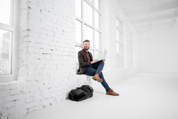 Elegante hombre de negocios sentado en la ventana con un portátil en el interior de la oficina blanca