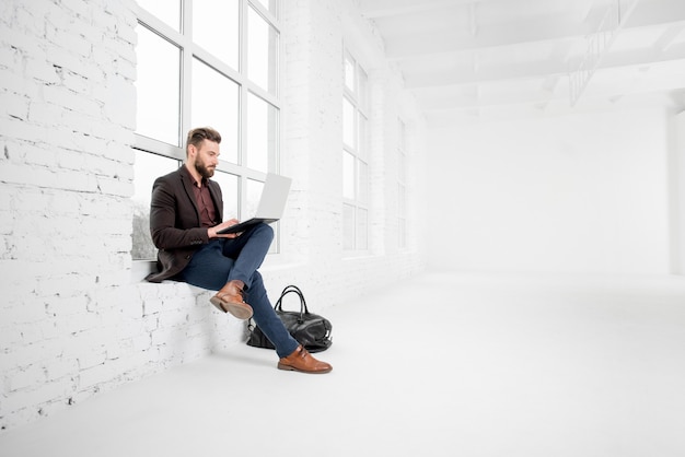 Elegante hombre de negocios sentado en la ventana con un portátil en el interior de la oficina blanca