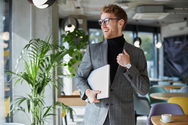 Elegante hombre de negocios con traje formal y anteojos sonríe y sostiene una computadora portátil en las manos en el interior de la oficina