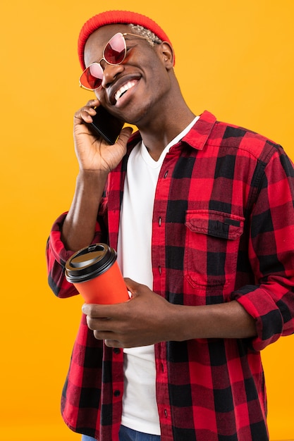 Elegante hombre negro americano con una hermosa sonrisa en una camisa roja a cuadros tiene en sus manos un vaso de café sobre un fondo amarillo studio