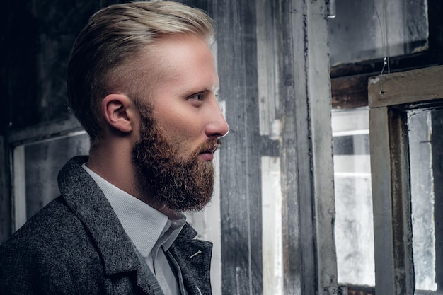 Elegante hombre rubio y barbudo vestido con una chaqueta elegante y clásica que mira a través del fondo de ventanas antiguas y antiguas.