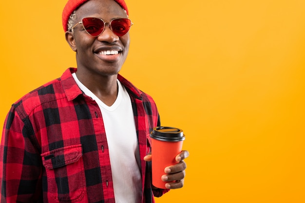 Elegante homem americano preto com um sorriso lindo em uma camisa quadriculada vermelha tem nas mãos um copo de café amarelo