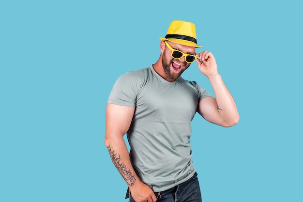 Elegante homem barbudo e musculoso e emocional com chapéu de palha amarelo, posando no estúdio sobre fundo azul