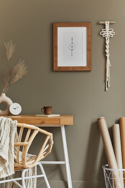 Elegante interior bohemio del espacio de la oficina en casa con escritorio de madera, sillón de ratán, marco de cartel marrón, macramé, suministros de oficina, decoración de la lámpara y elegantes accesorios personales en la decoración del hogar