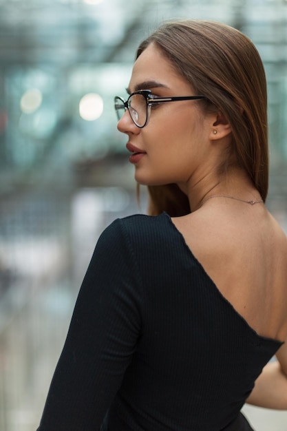 Elegante jovem bonita com óculos da moda em um top preto moderno com as costas abertas em um prédio de escritórios